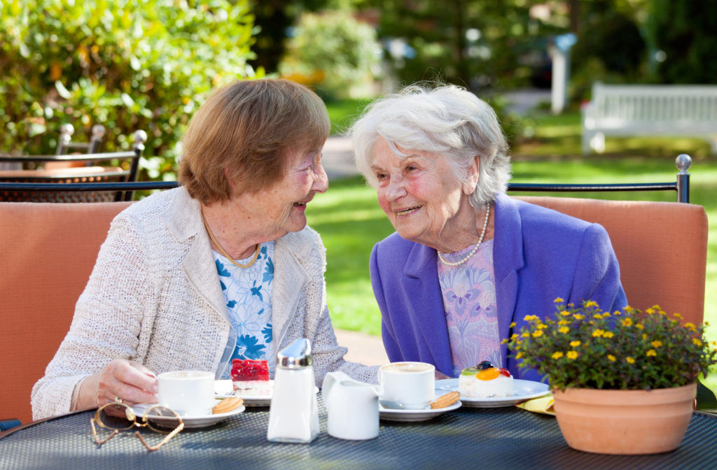 Two happy senior women talking to one another outside in backyard of deck sitting on patio