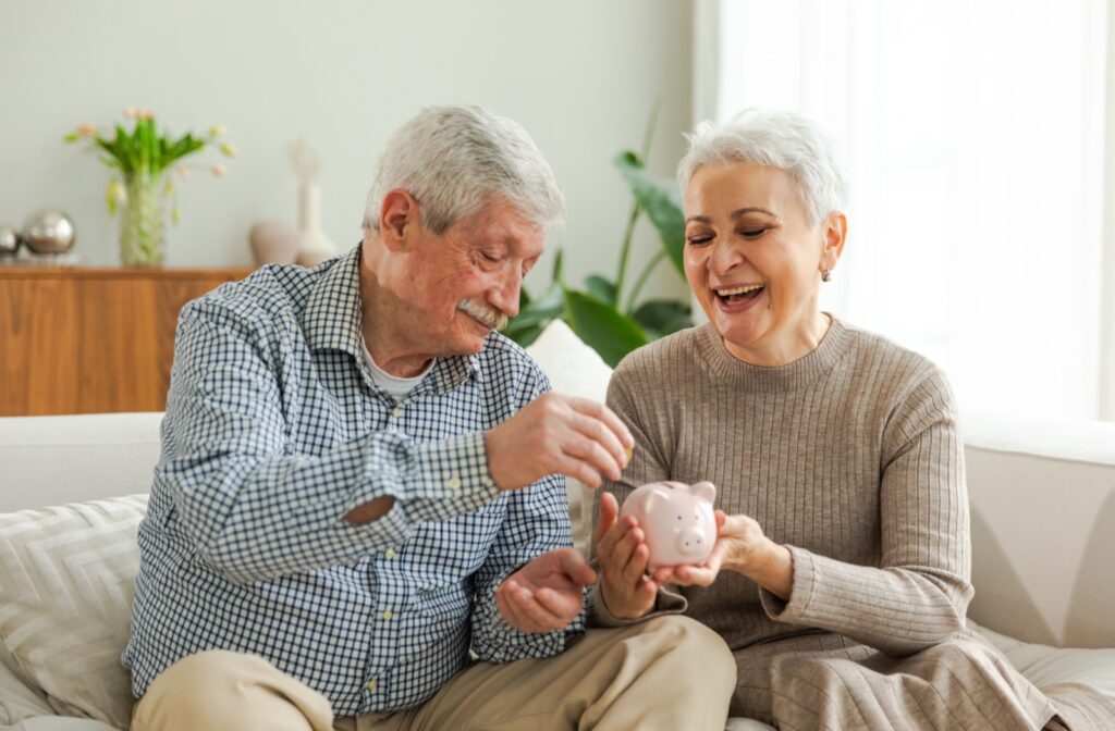 A senior couple holding a piggy bank in hand while the man places a dollar in it.