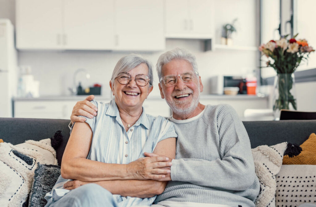 An older couple sitting in independent living, smiling with their arms around one another.