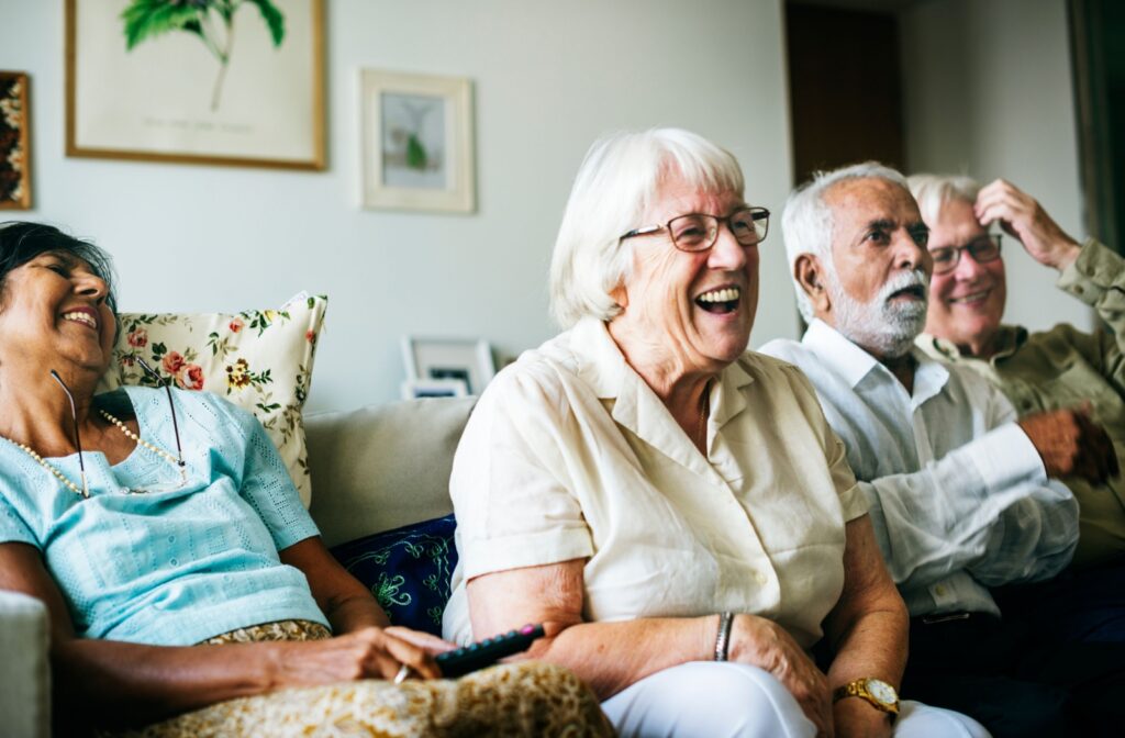 A group of older adults in senior living laugh while sitting on a couch in a resident apartment and watching a movie