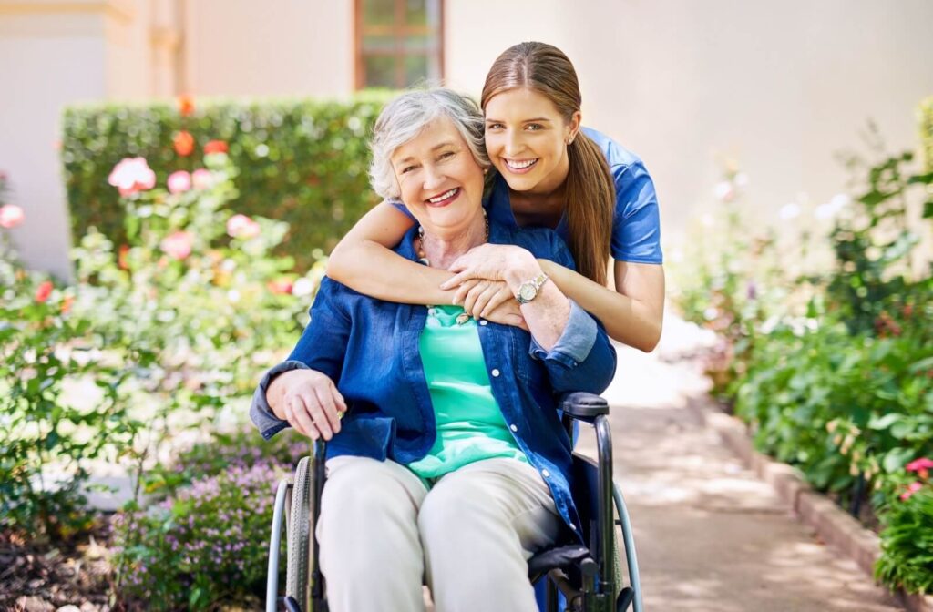 A caregiver hugs a resident from behind while rolling a wheelchair through beautiful community gardens